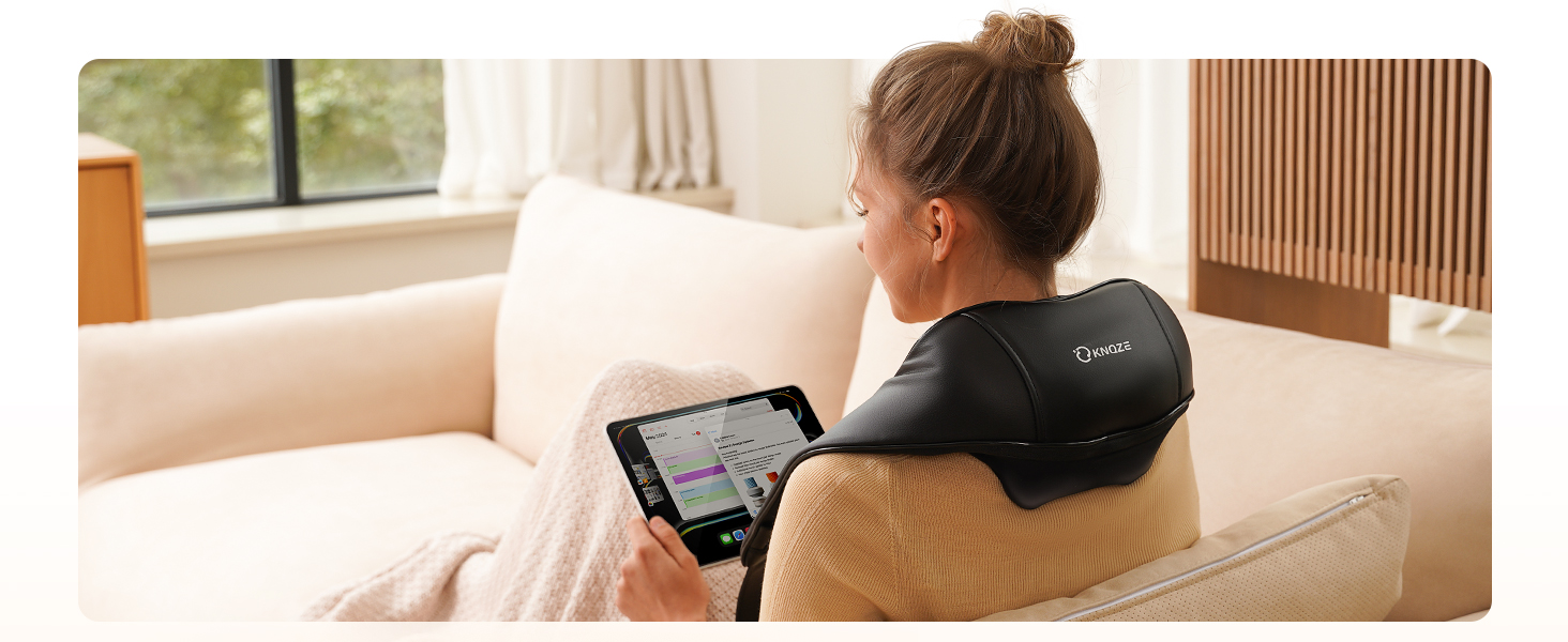 Woman using tablet while wearing neck massager on couch. Device has 'Comfier' branding, suggesting electric massage functionality for relaxation or pain relief.