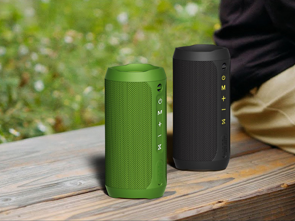 Two cylindrical portable speakers on wooden surface outdoors. Green speaker in foreground, black speaker behind. Both have mesh grilles and visible control buttons.