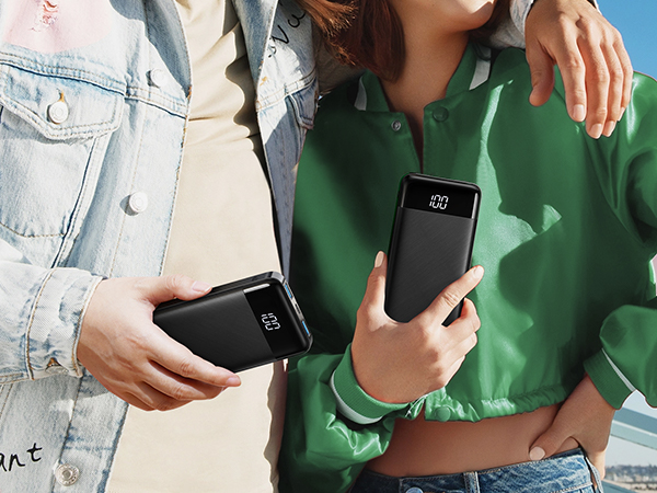 Two hands holding black portable power banks with digital displays showing battery percentage. Devices appear slim and rectangular in shape.