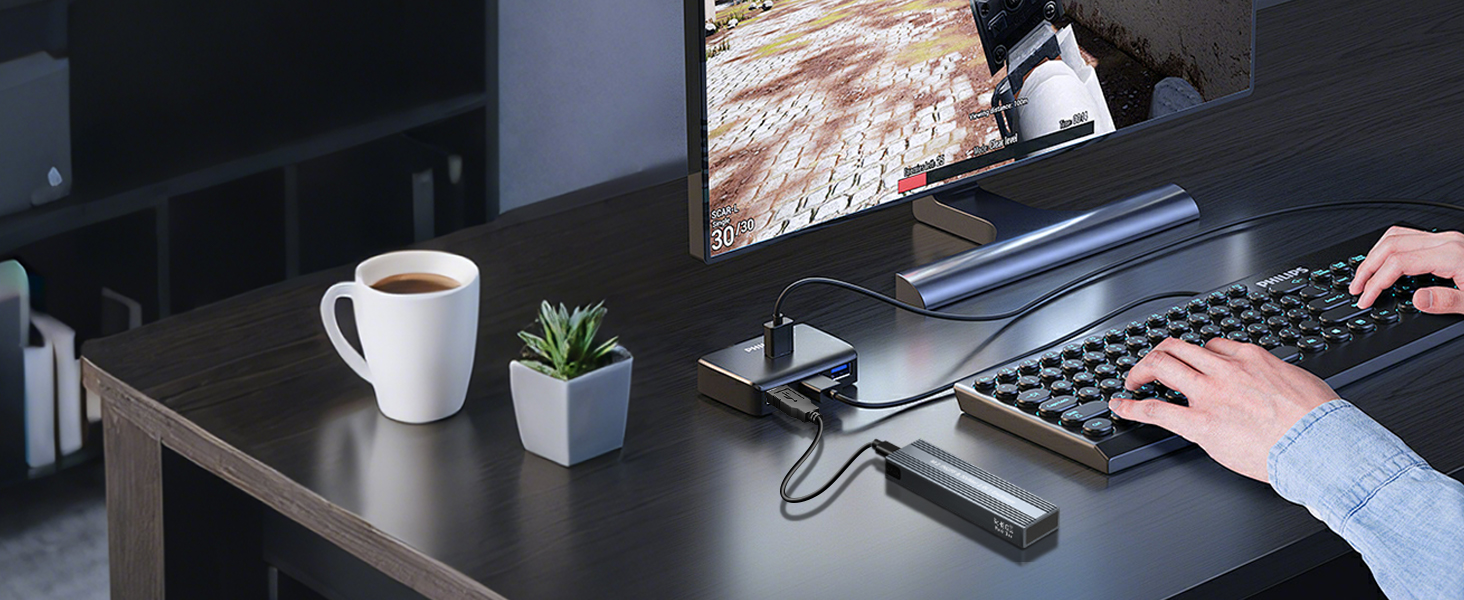 Desktop workspace with computer monitor, keyboard, and hands typing. Desk accessories include a white coffee mug and small plant. Dark wood desk surface visible.