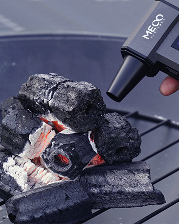 Black handheld device labeled 'MEEC' igniting charcoal briquettes. Glowing embers visible within the pile of charcoal on a grill grate.