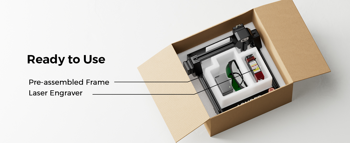 Opened cardboard box containing a pre-assembled laser engraver frame. The device has a black frame with silver components and a green circuit board visible.