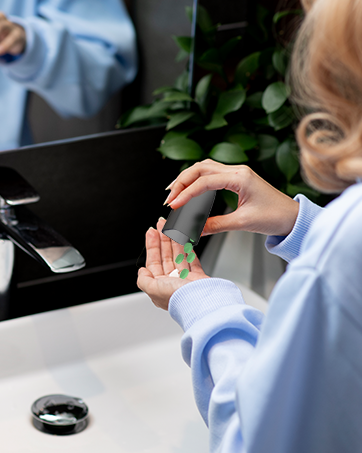 Hands dispensing liquid soap or sanitizer from a dark gray pump bottle in a bathroom setting with white countertop and green plants visible in background.