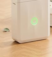 White rectangular air purifier or dehumidifier device with glowing green indicator light, placed on wooden flooring near a wall.