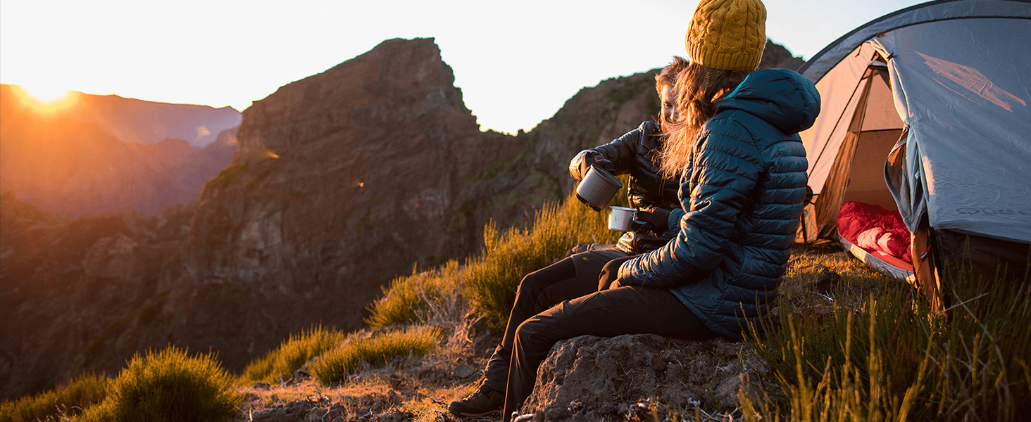 Camping scene with person sitting by tent, overlooking mountainous landscape at sunset. Gray tent pitched on grassy terrain near rocky peaks.