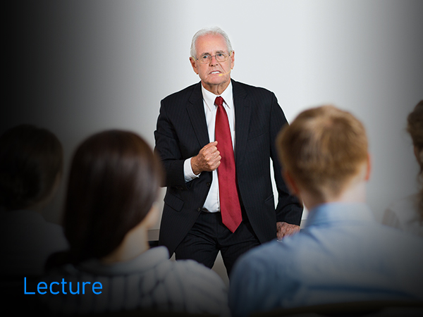 Professional presentation setting showing a speaker in formal business attire addressing an audience in a bright meeting space.