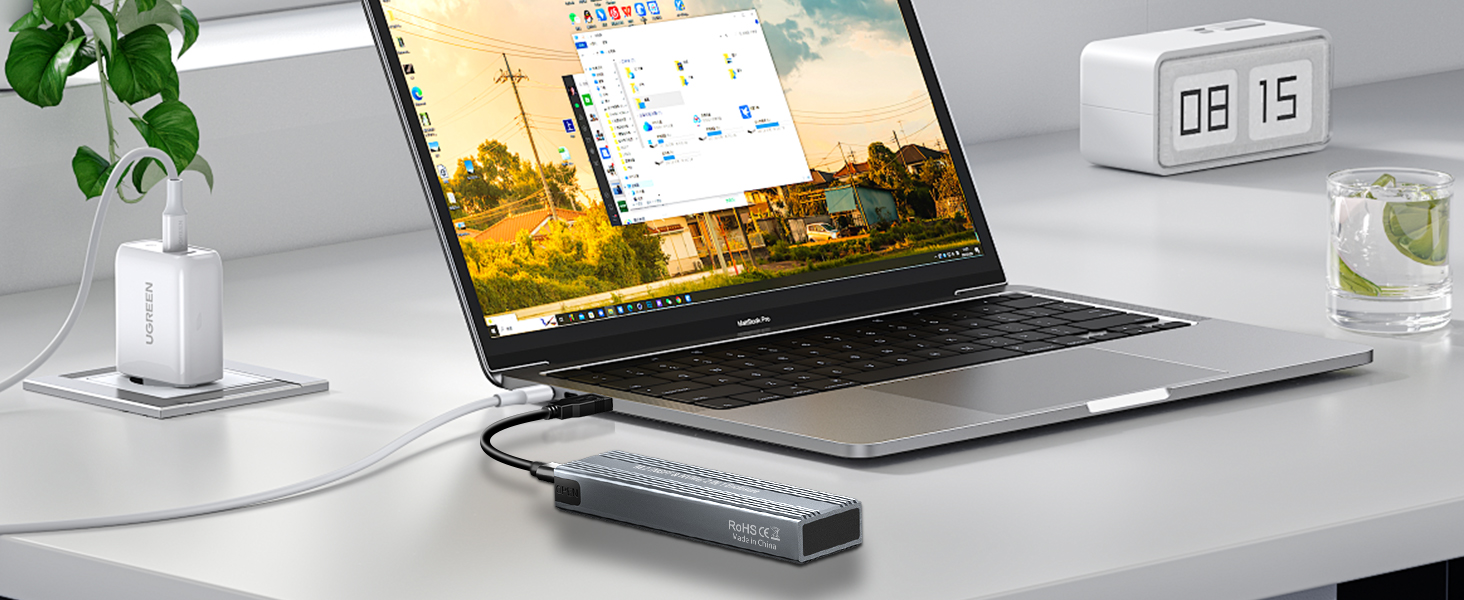 Slim laptop on desk with external device connected. Digital clock, plant, and glass nearby. Screen displays window with colorful icons and sunset background.