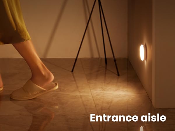 Illuminated motion sensor light on wall near floor, activating as person in slippers walks by. Warm glow on wooden floor, labeled 'Entrance aisle'.