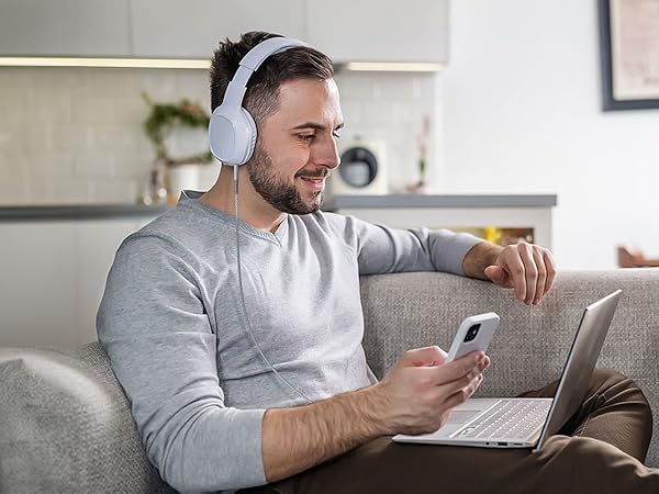 Person wearing white headphones and gray sweatshirt, seated on couch with laptop and mobile phone.