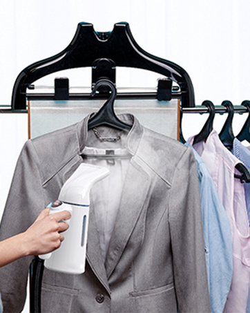 Handheld garment steamer in use on gray suit jacket, with multiple clothing items hanging on rack in background.