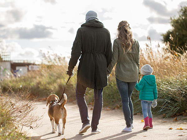 A family walking on a path with a dog. Adults in coats, child in teal jacket, and a brown dog on a leash in a natural setting with tall grass.