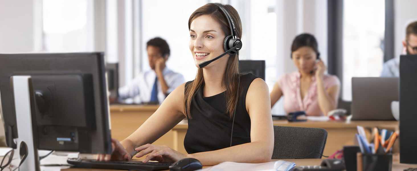 Customer service representatives working at computer workstations in an open office environment. Person wearing professional headset while operating computer at desk.