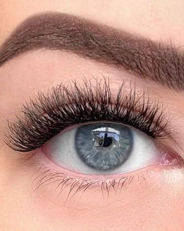 Close-up shot of eye wearing dramatic false eyelashes, showing full, voluminous black lashes against pale skin and a defined eyebrow.