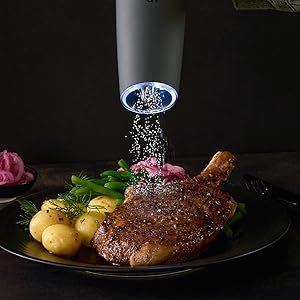 An image of the gravity grinder seasoning the steak with salt. Includes vegetable on a black plate
