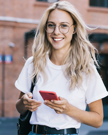 Person outdoors holding and looking at mobile phone while wearing casual white t-shirt and glasses.