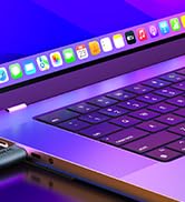 Partial view of a laptop keyboard and touchpad, illuminated by purple light. The laptop appears to be open with application icons visible on the screen.