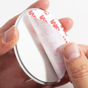 Hands holding a round, white compact mirror. The mirror's surface shows a partial reflection of red text, likely from packaging or labeling.