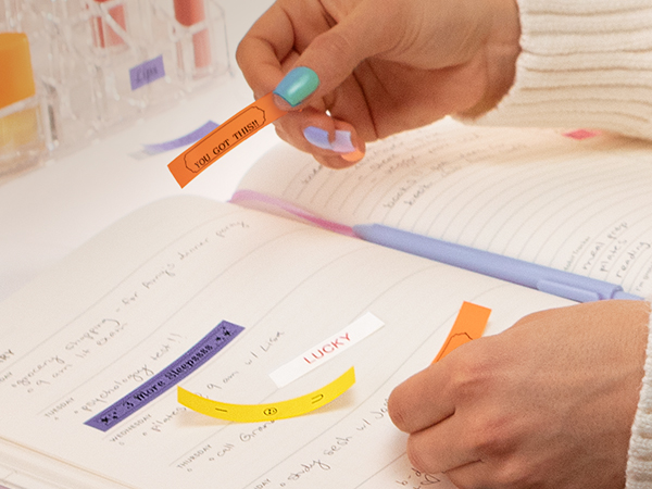 Person applying Brother P-touch Btag reminder and inspiration labels to planner notebook on desk