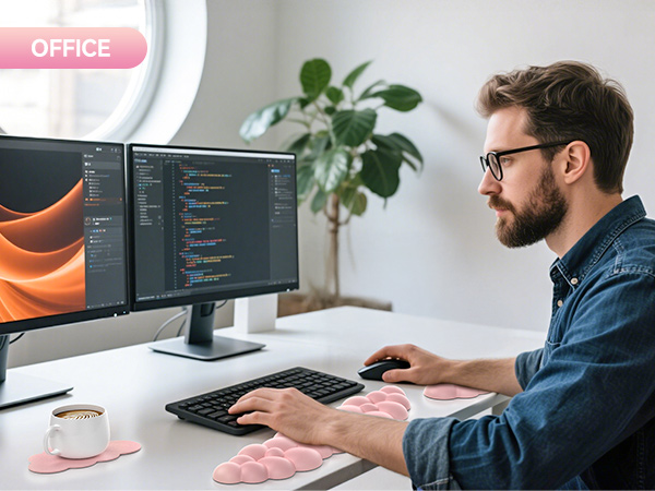 Developer working at desk with dual monitors displaying code. Ergonomic setup includes keyboard, mouse, wrist rest, and coffee cup on pink coaster. Potted plant in background.