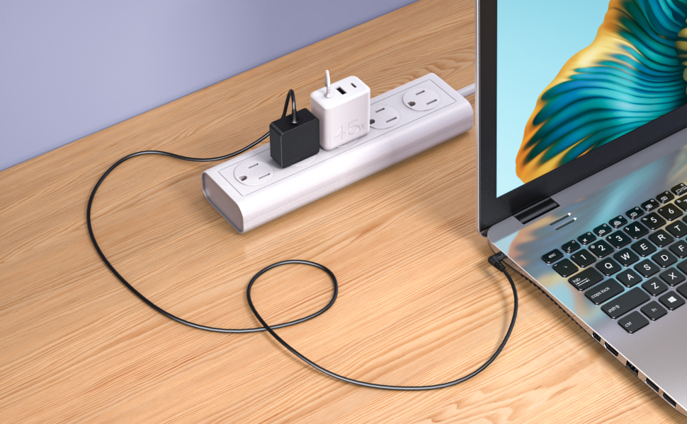A laptop connected to a power strip on a wooden desk with a neatly coiled charging cable.