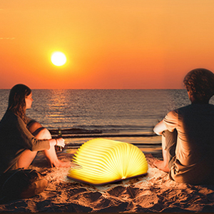 Glowing portable lamp on beach at sunset. Two silhouetted figures sit nearby. Lamp appears egg-shaped with warm yellow light.