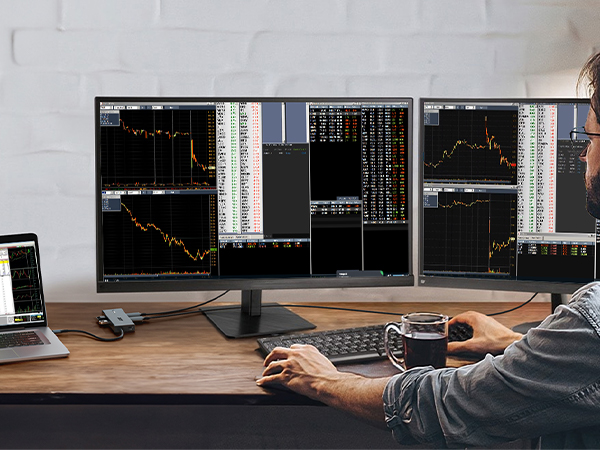 Trading setup with three monitors displaying financial charts and graphs, with keyboard and laptop on wooden desk.