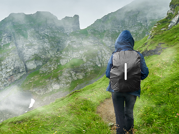 Hiker wearing grey and blue rain gear on misty mountain trail with steep green slopes and rocky peaks in background.
