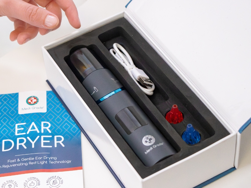 Text reads 'EAR DRYER'. Product packaging showcasing an electronic device in a black foam-lined case with blue accents and instructional icons.