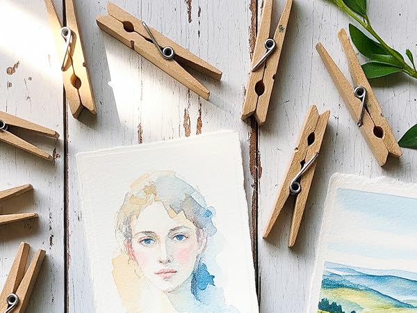 Wooden clothespins scattered around watercolor artwork on white surface. Two watercolor paintings visible - a portrait sketch and landscape scene.