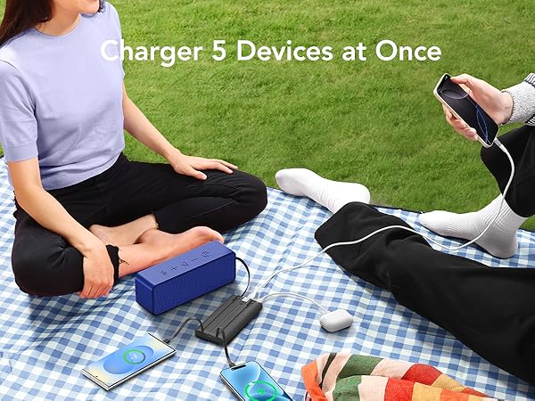 Outdoor picnic scene with people using a blue portable device capable of charging 5 devices simultaneously on a checkered blanket.