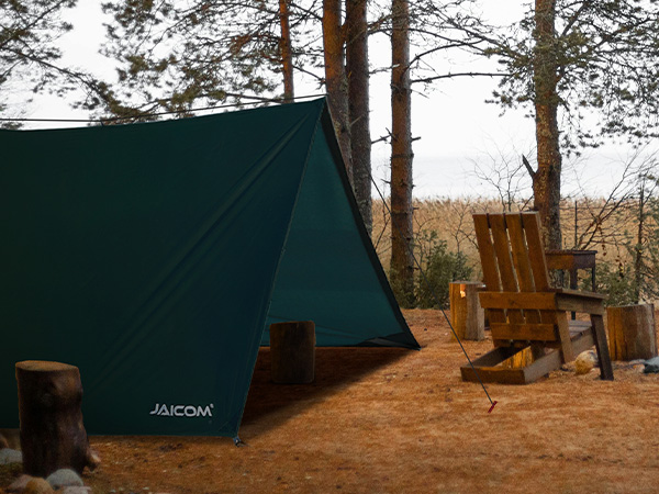 Green camping tent set up in a forest clearing. Two wooden chairs visible nearby, trees in background.
