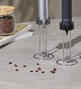 Two handheld milk frothers with metal whisks, one silver and one black, next to coffee beans and a jar. Close-up view on a gray surface.