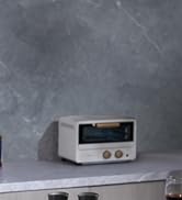 Compact white toaster oven with minimalist design featuring two control dials and a small viewing window on front panel, placed on light surface.