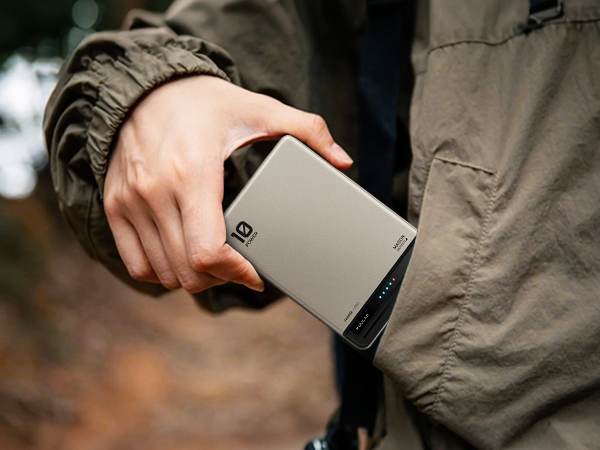 Hand holding a slim, silver portable power bank with black accents. Device features a digital display and multiple ports, indicating high-capacity external battery charger for mobile devices.