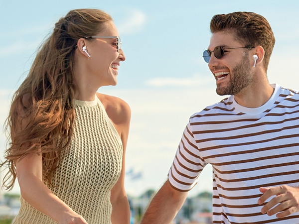 Two people wearing wireless earbuds outdoors, smiling in casual summer attire against a bright sky background.