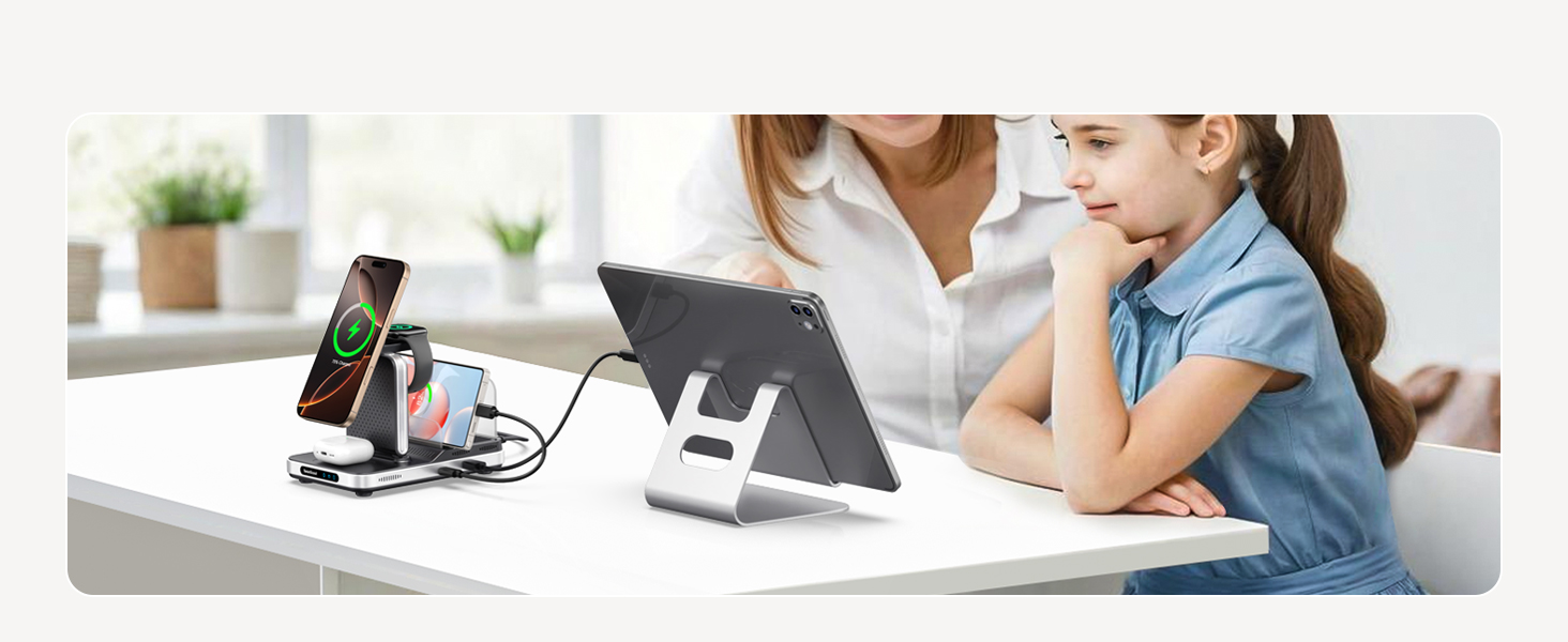 Charging station on desk with multiple device slots. Shows smartphone displaying battery icon, tablet stand, and USB ports. Two people visible in background.