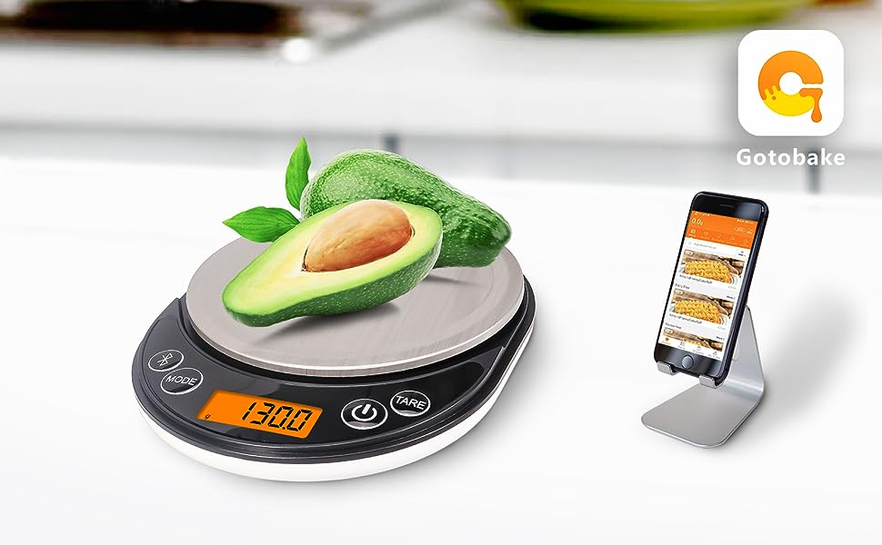 Digital kitchen scale with circular platform, displaying 1300g. Avocado on scale. Smartphone on stand nearby showing app interface.