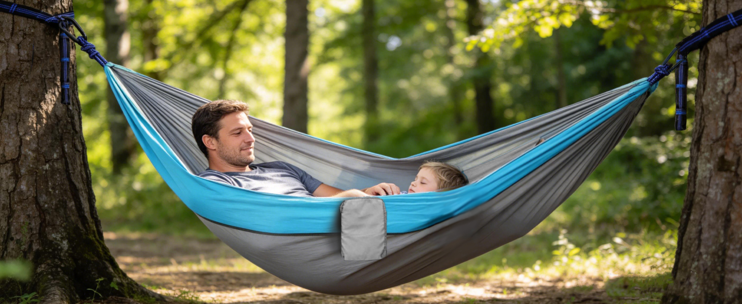 Person relaxing in a blue and gray camping hammock suspended between two trees in a sunny outdoor setting.