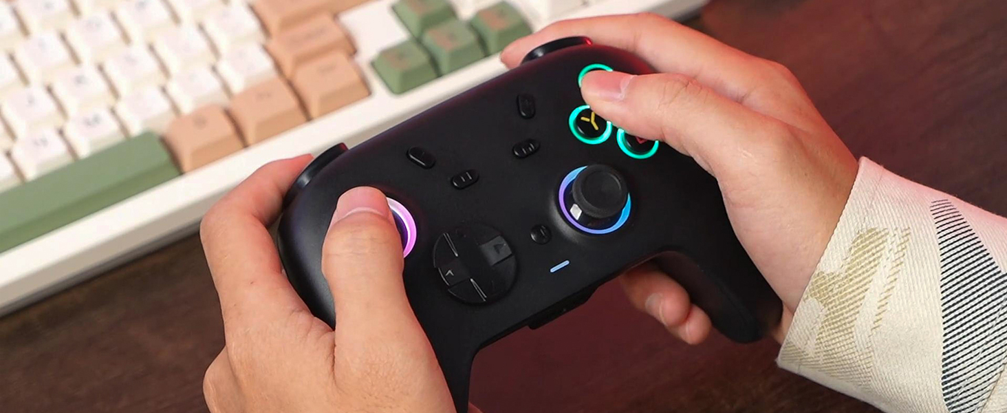 Hands holding a black video game controller with colorful illuminated buttons. The controller is being used above a partially visible keyboard with white and green keys.