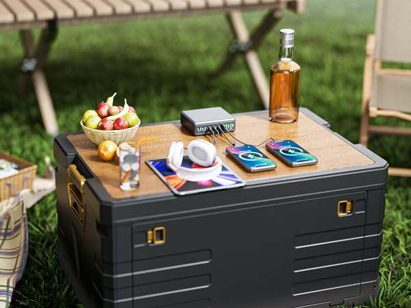 Outdoor scene with dark storage trunk used as table, holding phones, drinks, and fruit bowl on grass with natural daylight.
