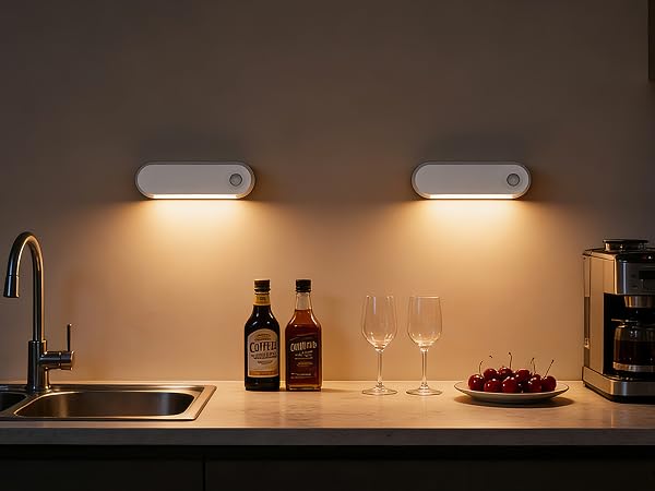Kitchen counter scene with two mounted light fixtures illuminating workspace. Features sink, bottles, wine glasses, and coffee maker in warm lighting.