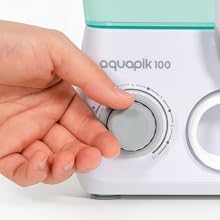 Text reads 'aquoptik 100'. Close-up of a hand turning a white control dial on a device with minimalist design featuring circular controls.