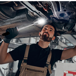 Mechanic in dark clothing and work apron examining vehicle undercarriage using handheld light while working underneath lifted car.
