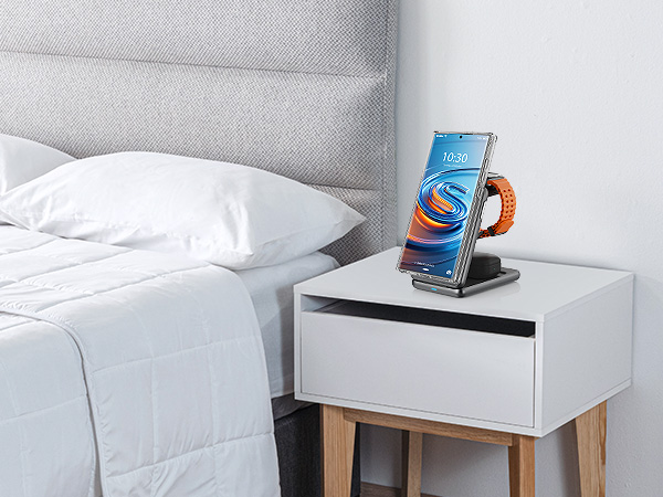 Wireless charging stand on bedside table, holding smartphone with orange wristband. Stand has angled design for easy viewing while charging.