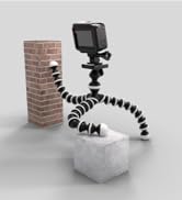 Flexible tripod with segmented legs wrapped around a brick column and concrete block, supporting a small action camera on top.