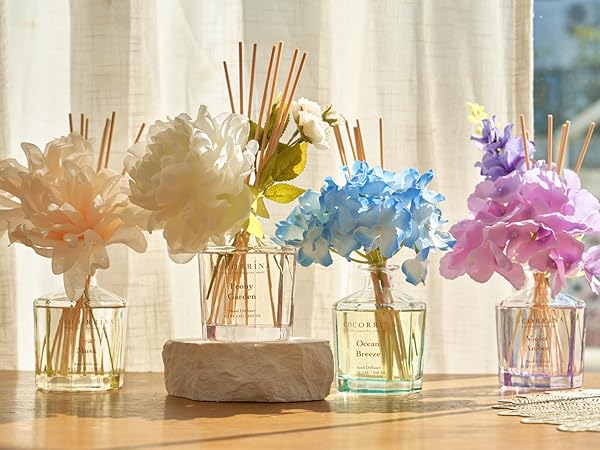 Four glass vases with colorful artificial flowers and reed diffusers, arranged on a wooden surface near a window with sheer curtains.
