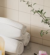 Corner of a bathroom with white tiled walls, folded white towels on a surface, and a green plant visible in the frame.