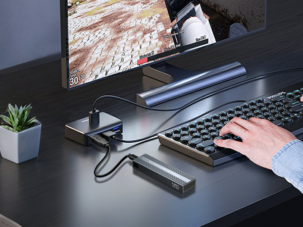 Desktop setup with curved monitor, keyboard, and docking station. Person's hand on keyboard. Small plant in white pot on desk. Dark surface with silver accessories visible.