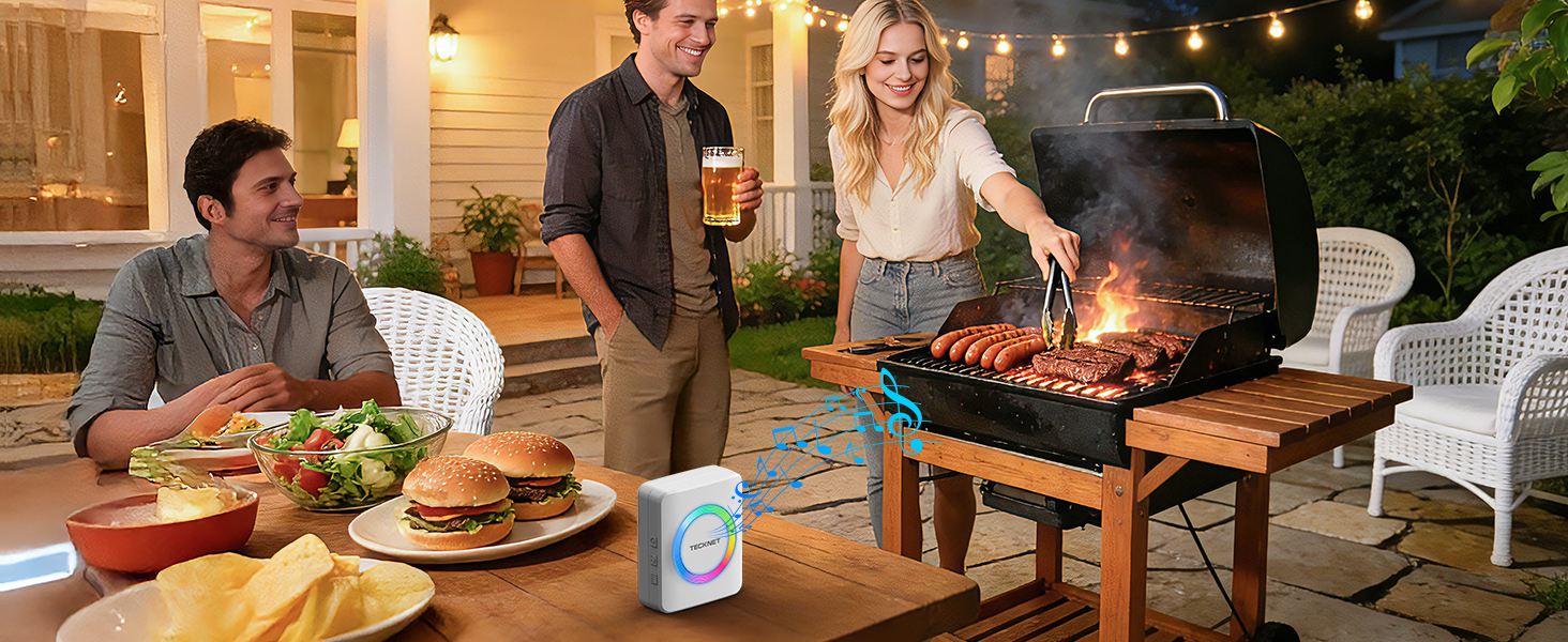 Outdoor evening scene with people gathered around a patio table for a barbecue dinner. String lights visible overhead creating ambient lighting.