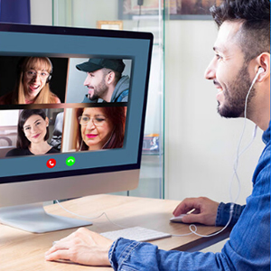 Desktop computer displaying video call with four participants. User engaged in conference, wearing earbuds, sitting at desk with keyboard.